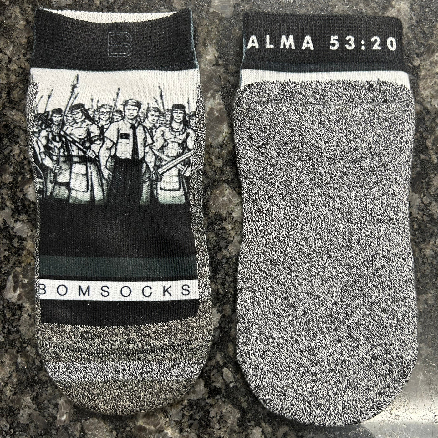 A pair of low-cut, gray and black socks with a black cuff featuring white text that reads 'ALMA 53:20'.